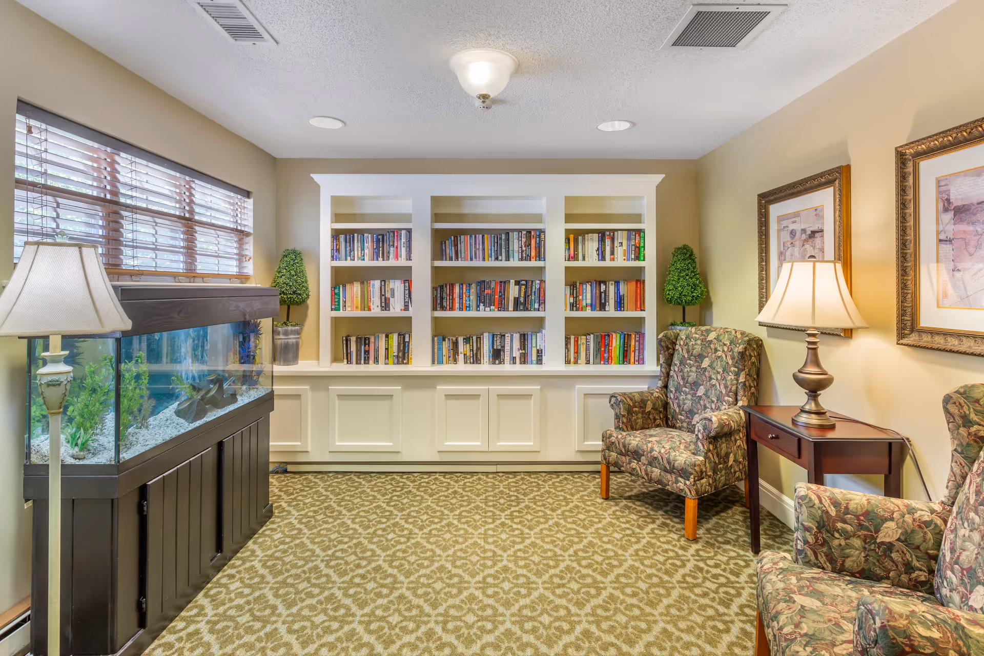 A cozy reading room with a large white built-in bookshelf filled with books, two floral upholstered armchairs, a wooden side table with a lamp, two framed pictures on the wall, a floor lamp, and a large fish tank beneath a window with wooden blinds.