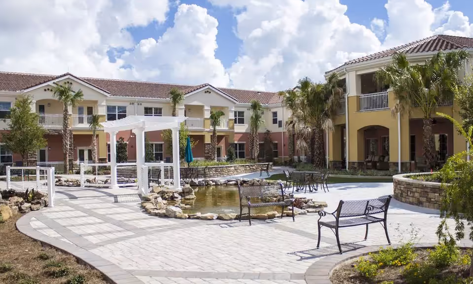 Courtyard of a senior living facility with a central pond, benches, a white pergola, and surrounding two-story buildings with balconies.