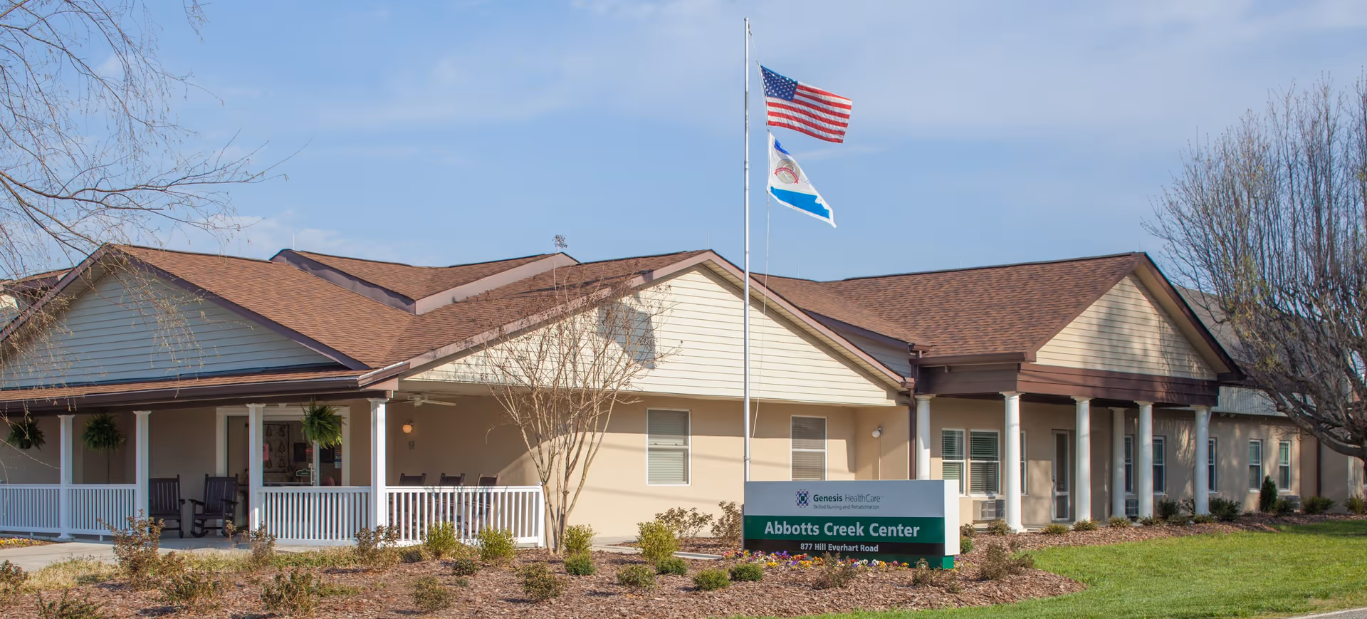 Exterior view of Abbotts Creek Center, a single-story building with a brown roof and beige walls. There is a covered porch with white railings and rocking chairs. Two flags, including an American flag, are flying on a flagpole in front of the building. The surrounding area has some landscaping with bushes and trees.