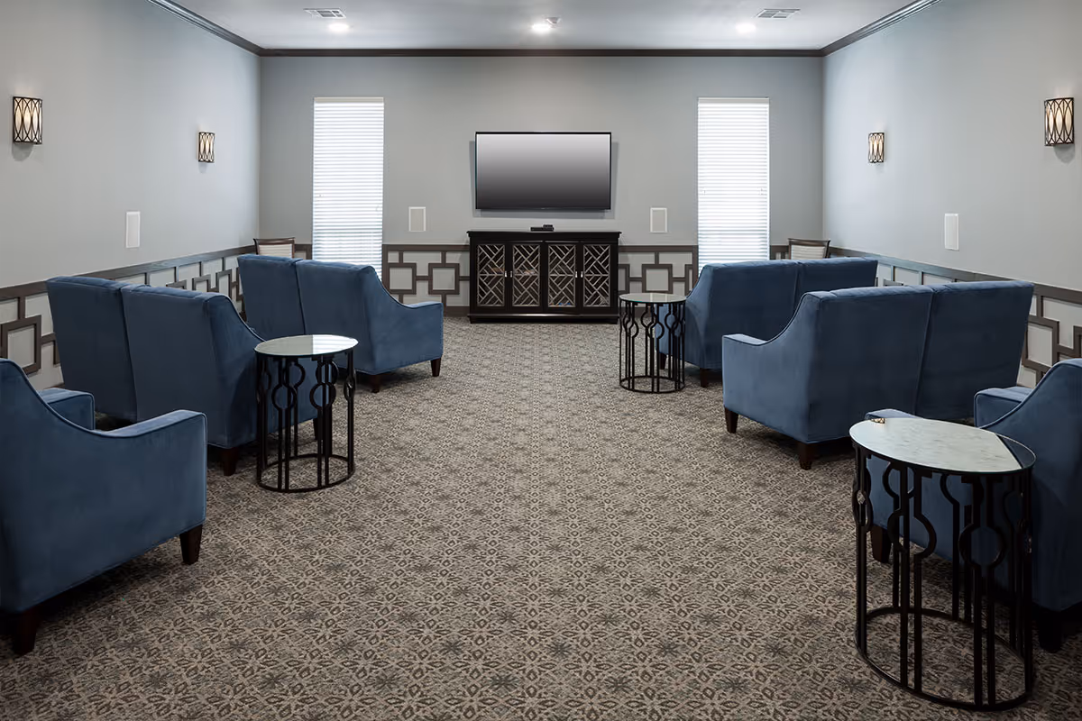 Carpeted lounge with blue upholstered armchairs arranged facing a wall-mounted TV and decorative side tables.