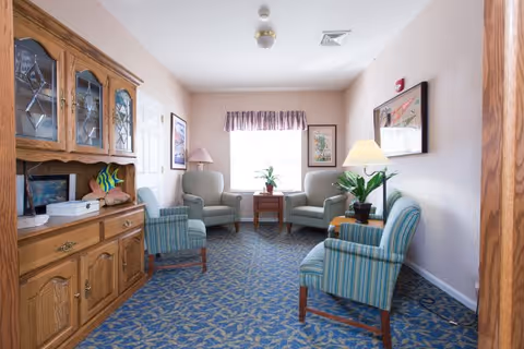 A small sitting room with four upholstered armchairs arranged around a wooden side table with a plant. The room has patterned blue carpet, light-colored walls with framed artwork, a wooden cabinet with glass doors on the left, and two lamps providing soft lighting.