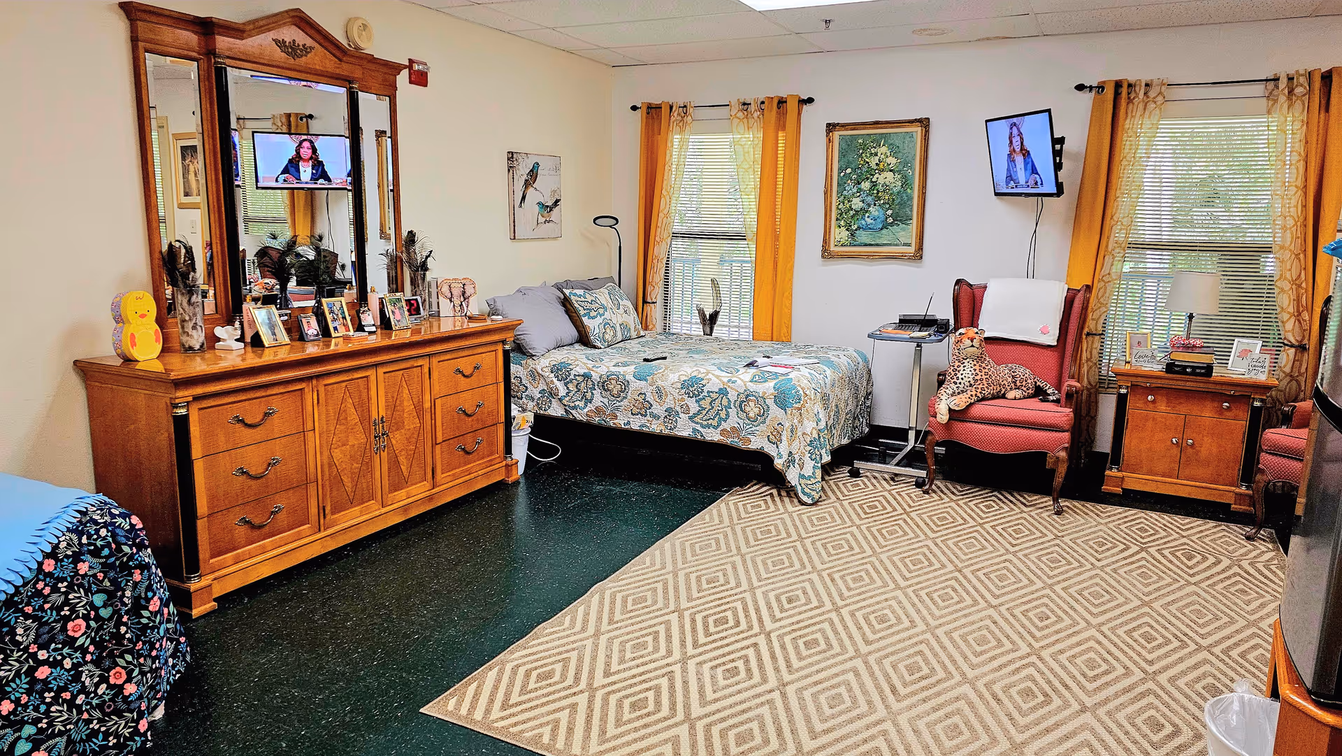 A cozy bedroom in an assisted living facility featuring a single bed with floral bedding, a wooden dresser with a large mirror and framed photos, two red armchairs with a stuffed leopard toy on one, a patterned area rug, two windows with yellow curtains, and two wall-mounted televisions.