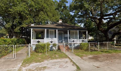 Front view of a small single-story residential care house with a porch and chain-link fence.