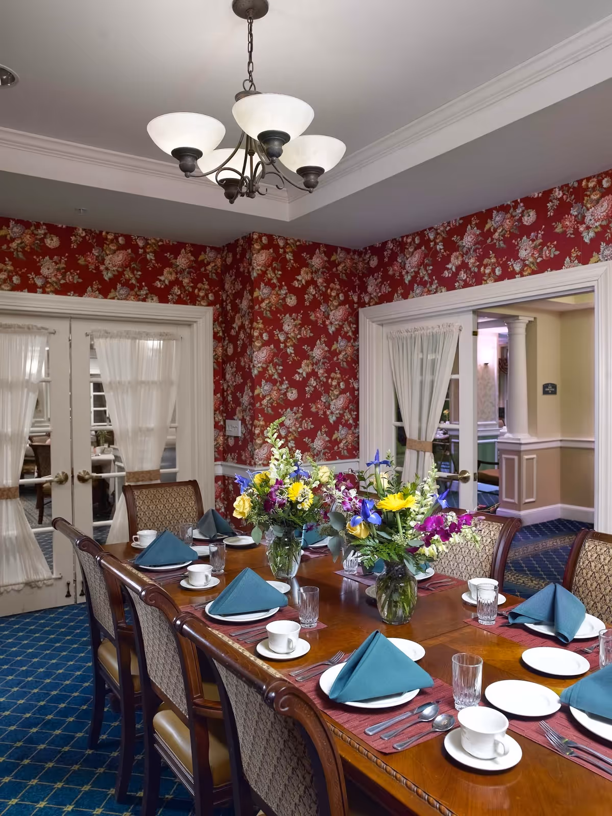 Formal dining table set with folded napkins, place settings and floral centerpieces in a room with red floral wallpaper.