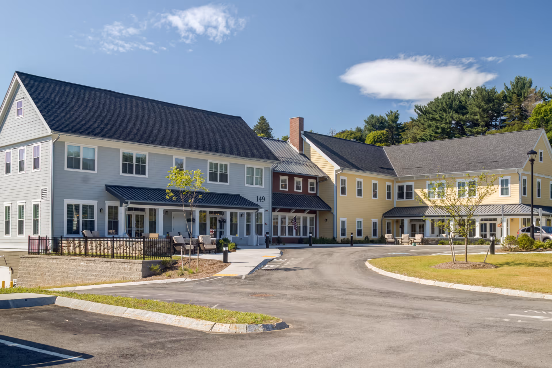Front exterior of a two-story senior living building with a circular driveway, patio seating, and landscaped grounds.
