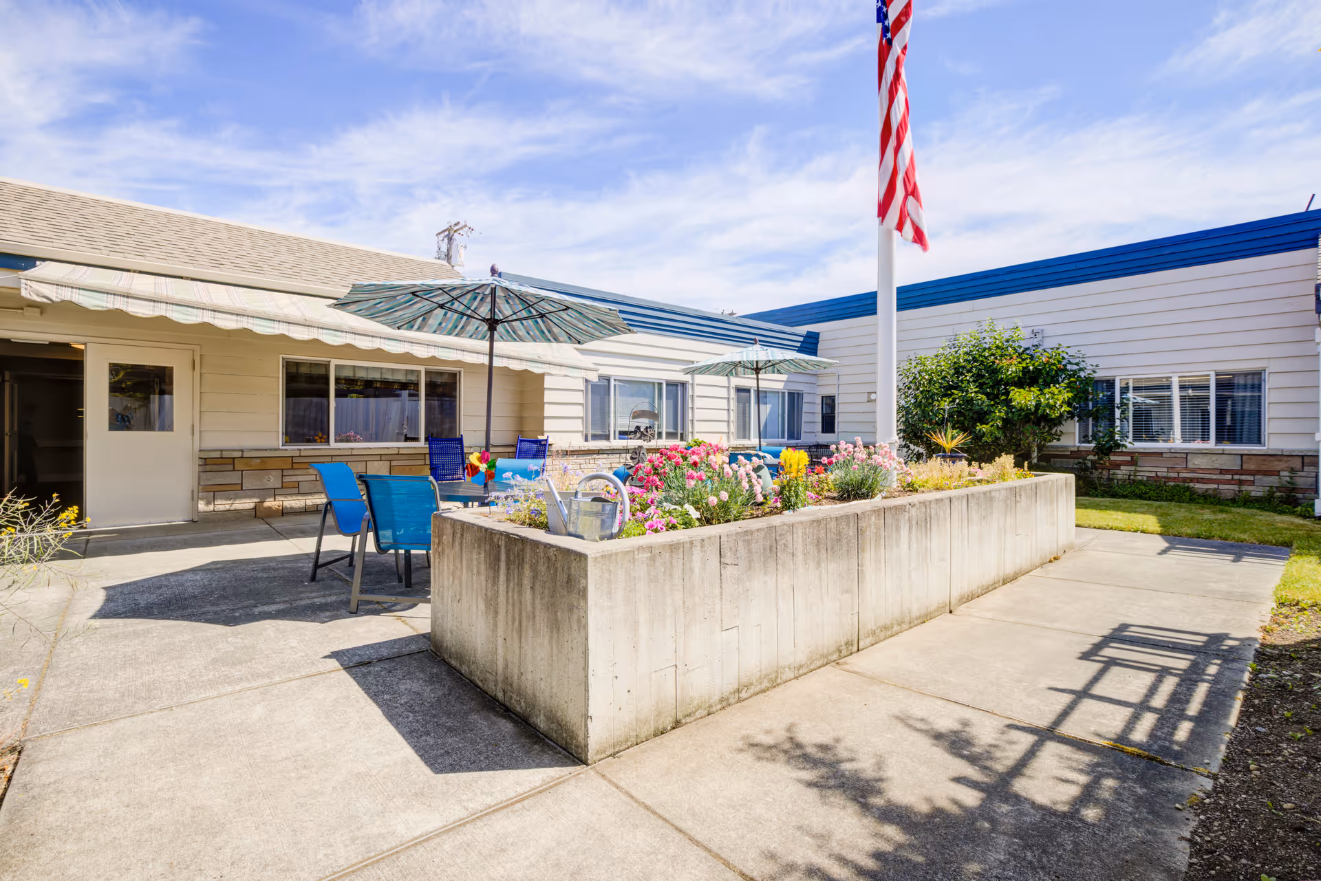 Outdoor patio area at The Cottages of Sequim featuring a concrete planter filled with colorful flowers, blue chairs, striped umbrellas, and an American flag on a flagpole. The building exterior is visible with white siding and windows under a partly cloudy sky.