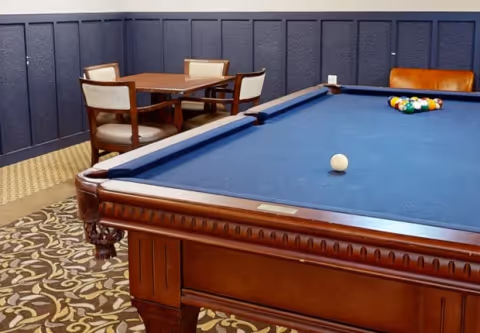 A blue-felt pool table in a carpeted game room with a small table and chairs against paneled walls.