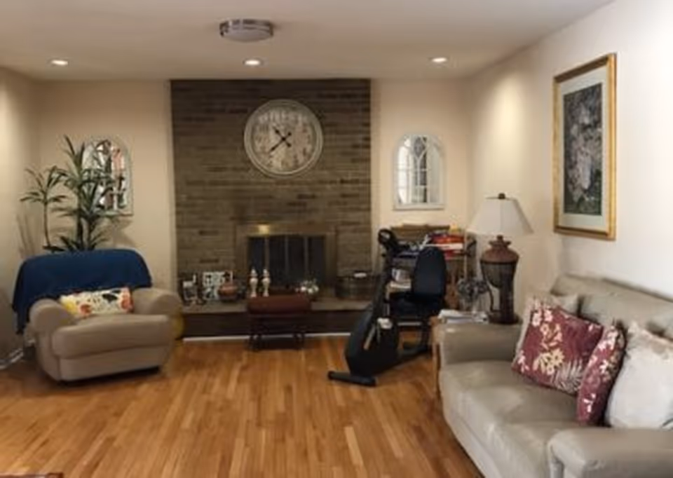 A cozy living room with wooden flooring, a beige sofa with floral cushions on the right, a beige armchair with a blue throw and a floral pillow on the left, a brick fireplace with a large round clock above it in the center, two small arched mirrors on either side of the fireplace, a black exercise bike, a wooden side table with a lamp and books, and a framed floral painting on the wall.
