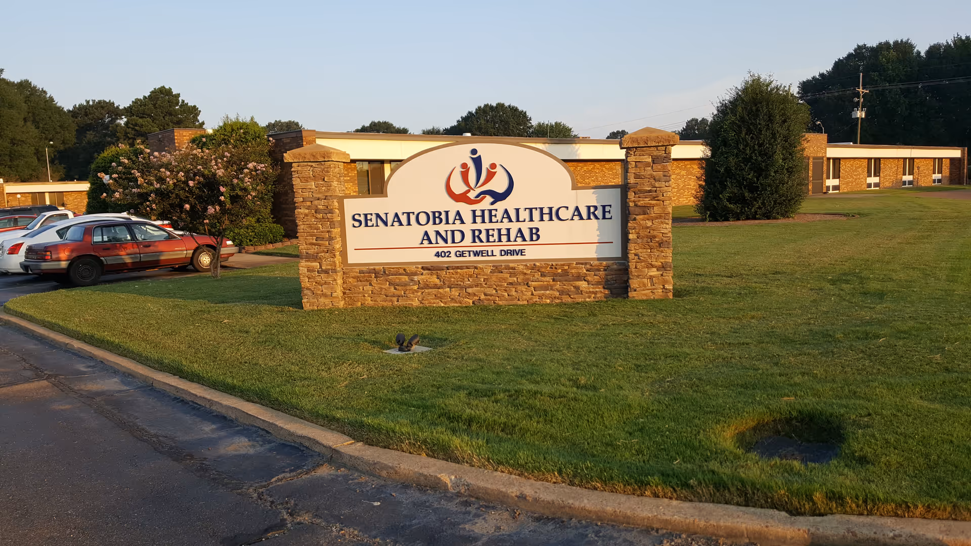 Outdoor view of Senatobia Healthcare and Rehab facility sign on a grassy area with a brick base, surrounded by trees and parked cars, with the building visible in the background under a clear sky.