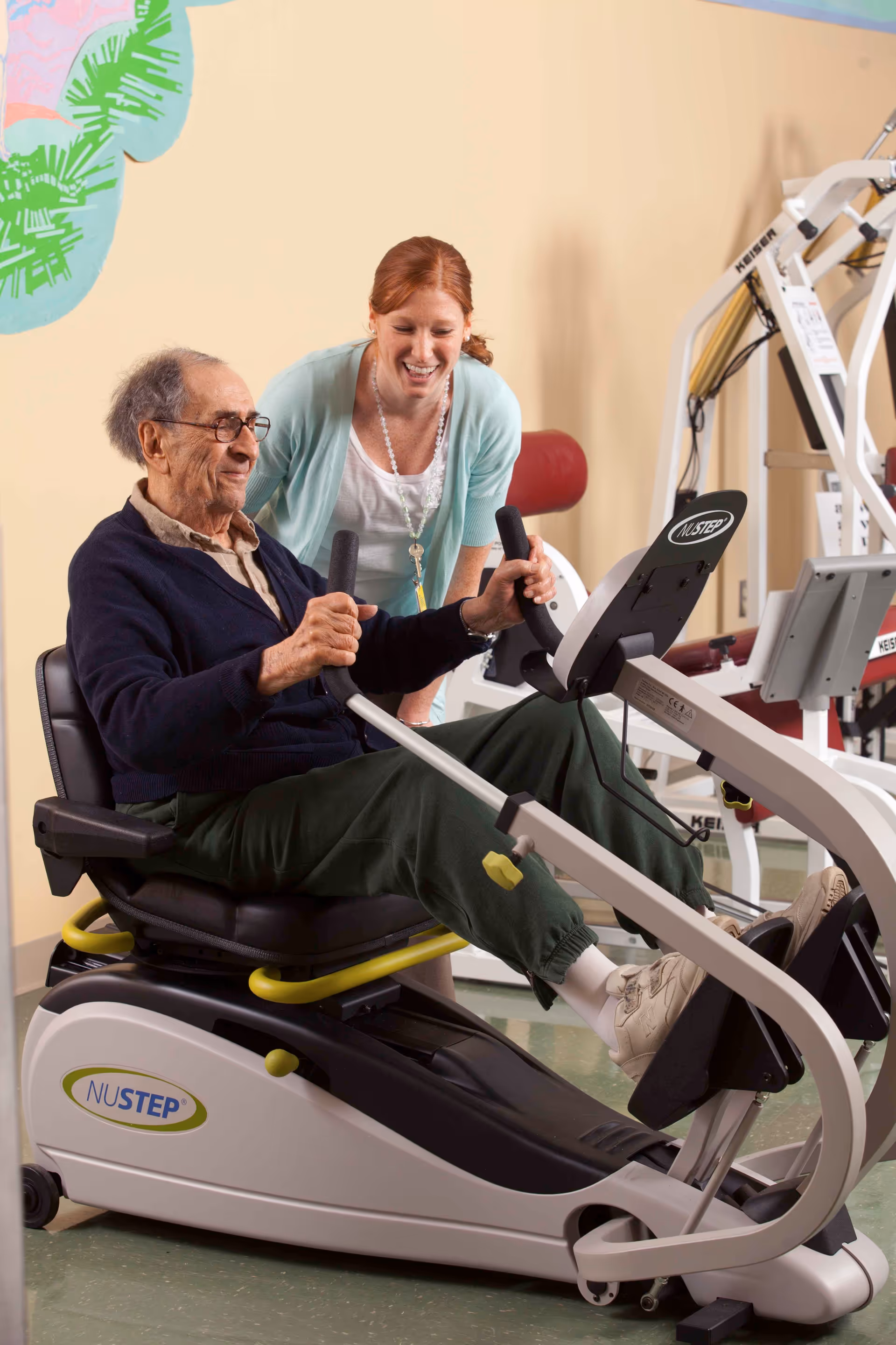 An elderly man is seated on a NuStep exercise machine, using the handles and pedals for a workout. A smiling woman, possibly a caregiver or physical therapist, is standing beside him, offering support and encouragement. The setting appears to be a rehabilitation or fitness room with exercise equipment in the background.