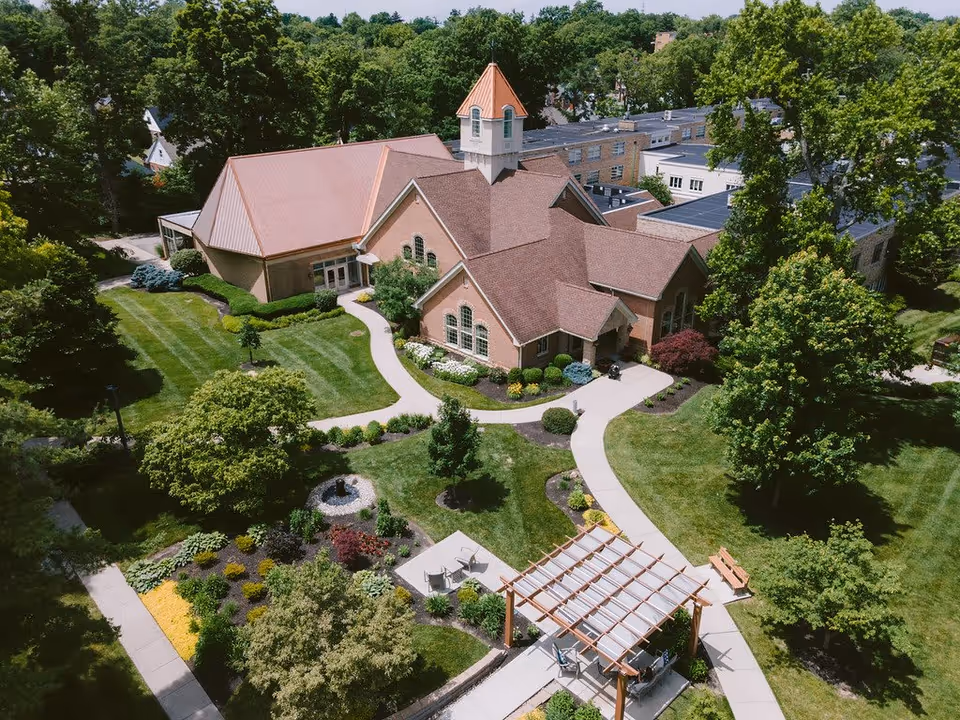 Aerial view of Ohio Living Llanfair facility showing a large brick building with a peaked roof and a small tower. The building is surrounded by well-maintained green lawns, trees, landscaped gardens, walking paths, and outdoor seating areas with pergolas and benches.