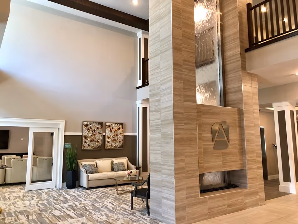 A spacious and modern lobby area with high ceilings, a large decorative water feature with the letter 'A' on it, a beige sofa with cushions, a glass coffee table, two framed floral artworks on the wall, a tall green plant in a black pot, and a seating area visible through glass doors.