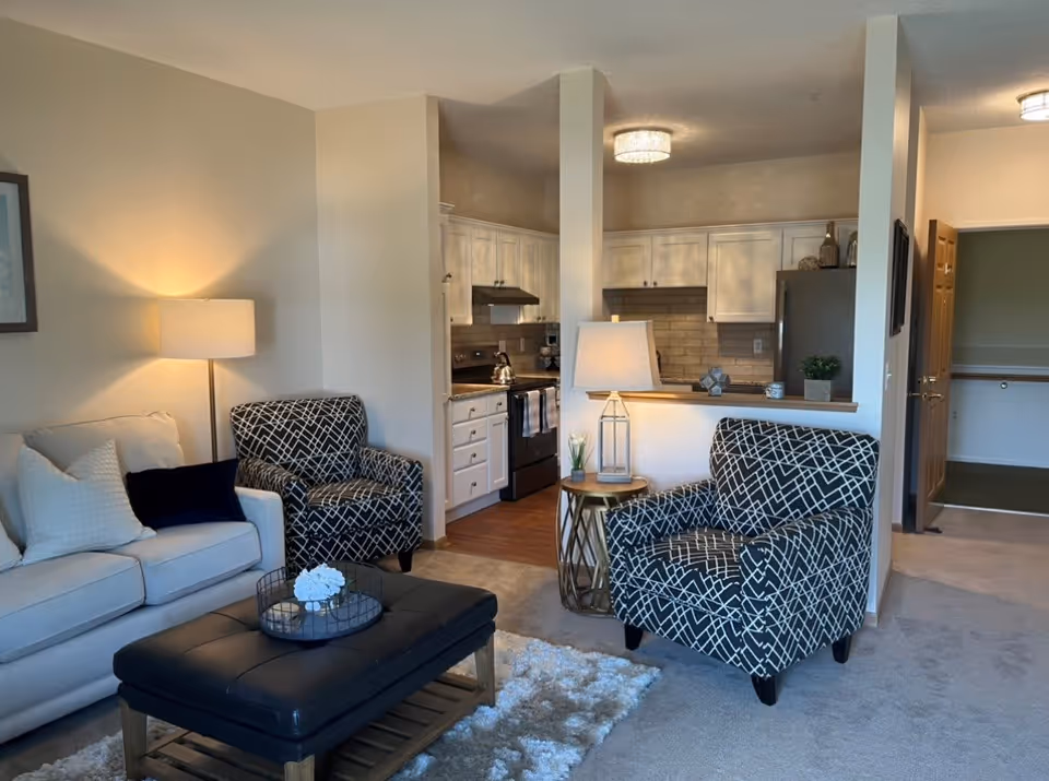 A cozy living room area with a beige sofa, two patterned armchairs, a black cushioned ottoman, and a side table with a lamp. The room opens into a kitchen with white cabinets and stainless steel appliances. The walls are painted light beige, and there is a soft carpet on the floor.