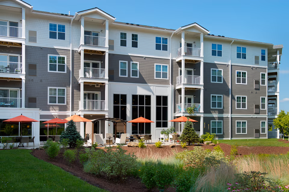 Exterior view of a multi-story senior living building with balconies, patio umbrellas, seating areas, and landscaped grounds.