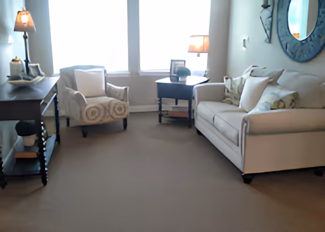 A cozy living room with a beige sofa adorned with cushions, a patterned armchair, a dark wooden side table with a lamp, and a console table with decorative items. A large round mirror hangs on the wall above the sofa, and bright natural light comes through the windows in the background.