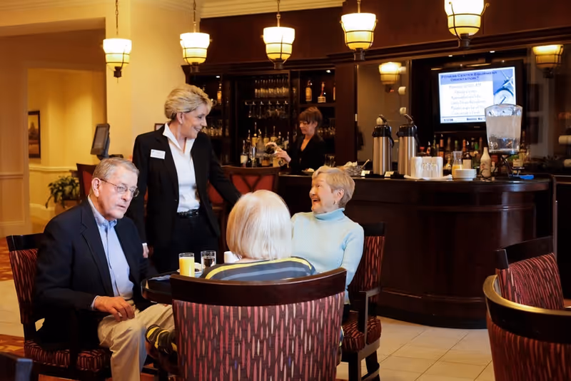 Three elderly people sitting around a table in a lounge area with drinks, talking and smiling, while a staff member stands nearby engaging with them. In the background, a woman is preparing drinks behind a bar counter with hanging lights above.