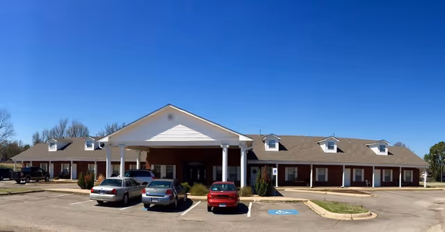 Front exterior view of Clarksville Retirement Center, a single-story brick building with a covered entrance supported by white columns. Several cars are parked in front under a clear blue sky.