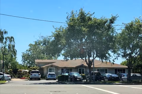 One-story building with several parked cars and large trees in a sunny parking lot.