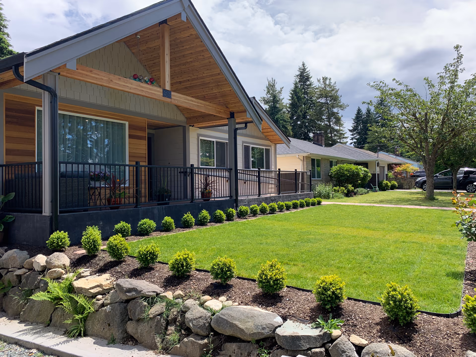 A single-story residential building with a covered porch and black railing, surrounded by a well-maintained lawn and small shrubs. There are trees and other houses visible in the background under a partly cloudy sky.