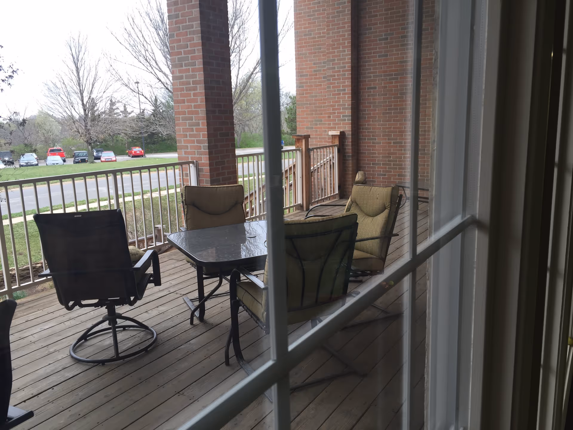 View through a window onto a covered wooden deck with a glass-top patio table, several cushioned chairs, brick columns, and a railing overlooking a parking area.