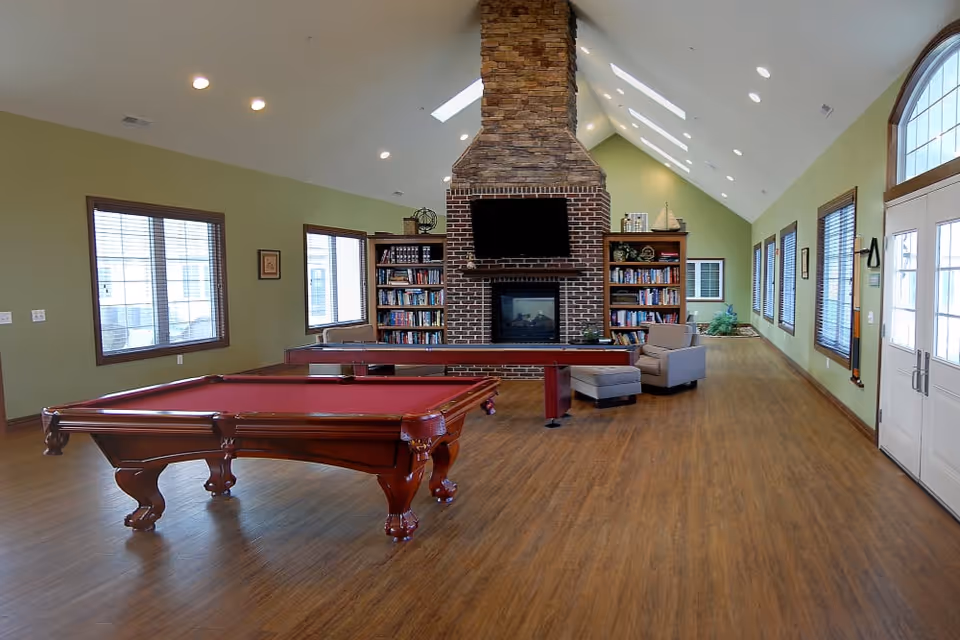 A spacious recreational room with a red pool table in the foreground, a shuffleboard table behind it, and a large brick fireplace with a mounted TV above it. The room has wooden floors, green walls, multiple windows with blinds, bookshelves filled with books, and comfortable seating including an armchair with an ottoman. Skylights and recessed lighting brighten the vaulted ceiling.