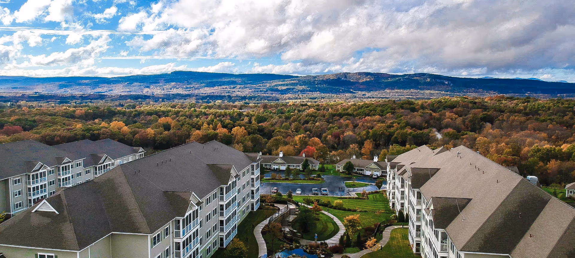 Aerial view of Woodland Pond At New Paltz senior living facility showing multiple large residential buildings with gray roofs surrounded by green lawns, trees with autumn foliage, and a winding pathway. In the background, there are forested hills under a partly cloudy blue sky.