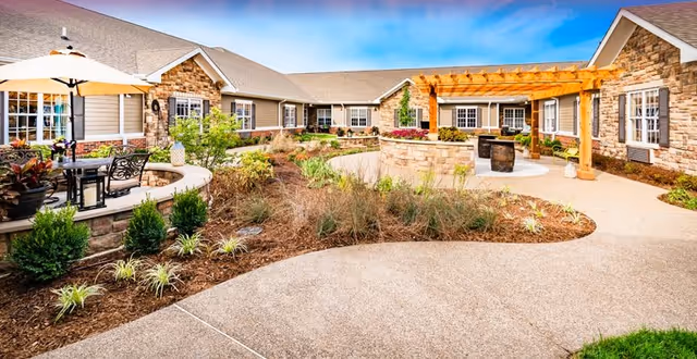Sunlit outdoor courtyard of a senior living facility with stone-clad buildings, landscaped beds, patio seating, and a wooden pergola.