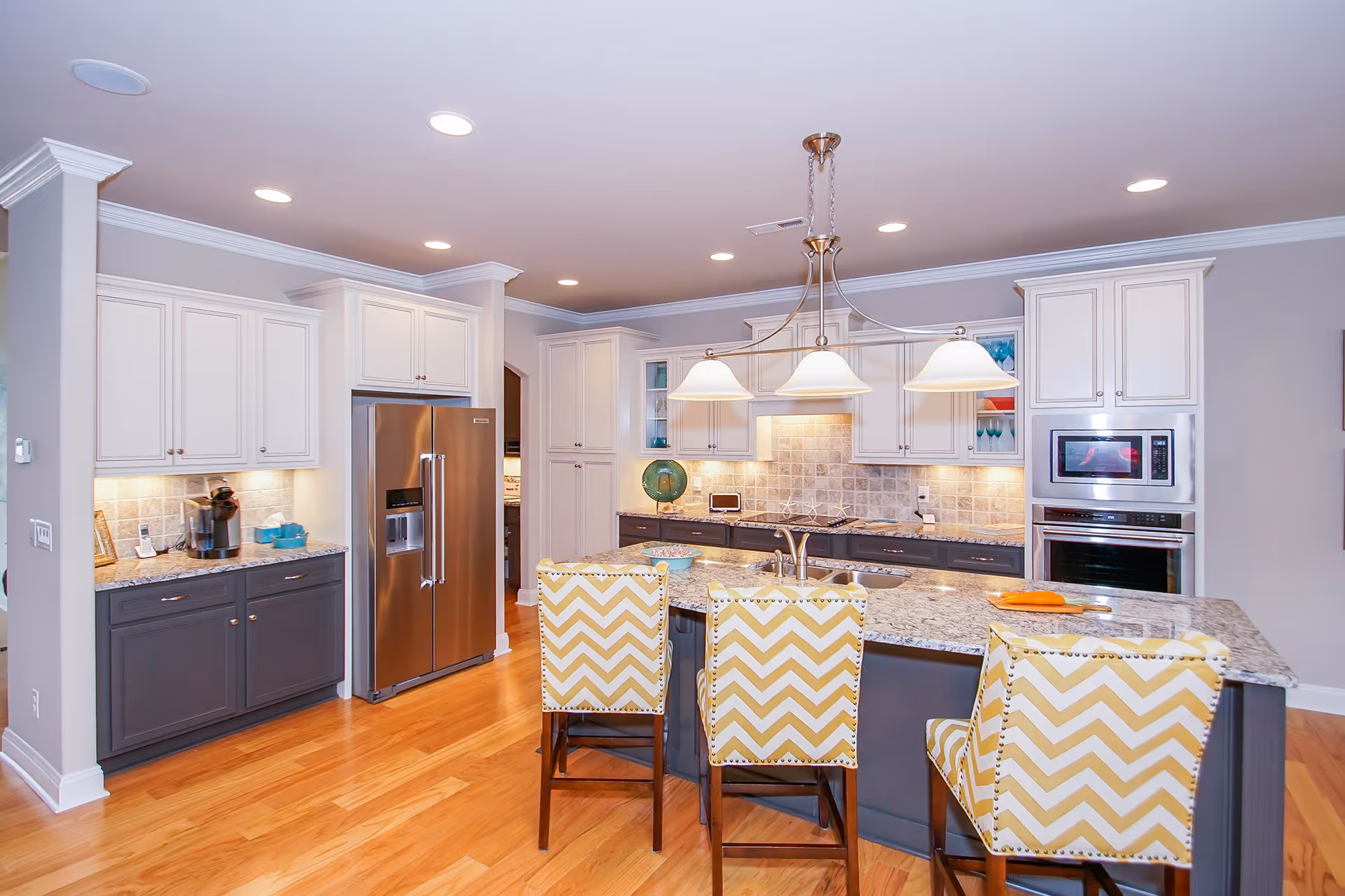 Bright modern kitchen with white and dark gray cabinets, stainless steel refrigerator, built-in microwave and oven, granite countertops, and a kitchen island with three yellow and white chevron-patterned chairs. The kitchen has wooden flooring and recessed lighting with a hanging light fixture above the island.