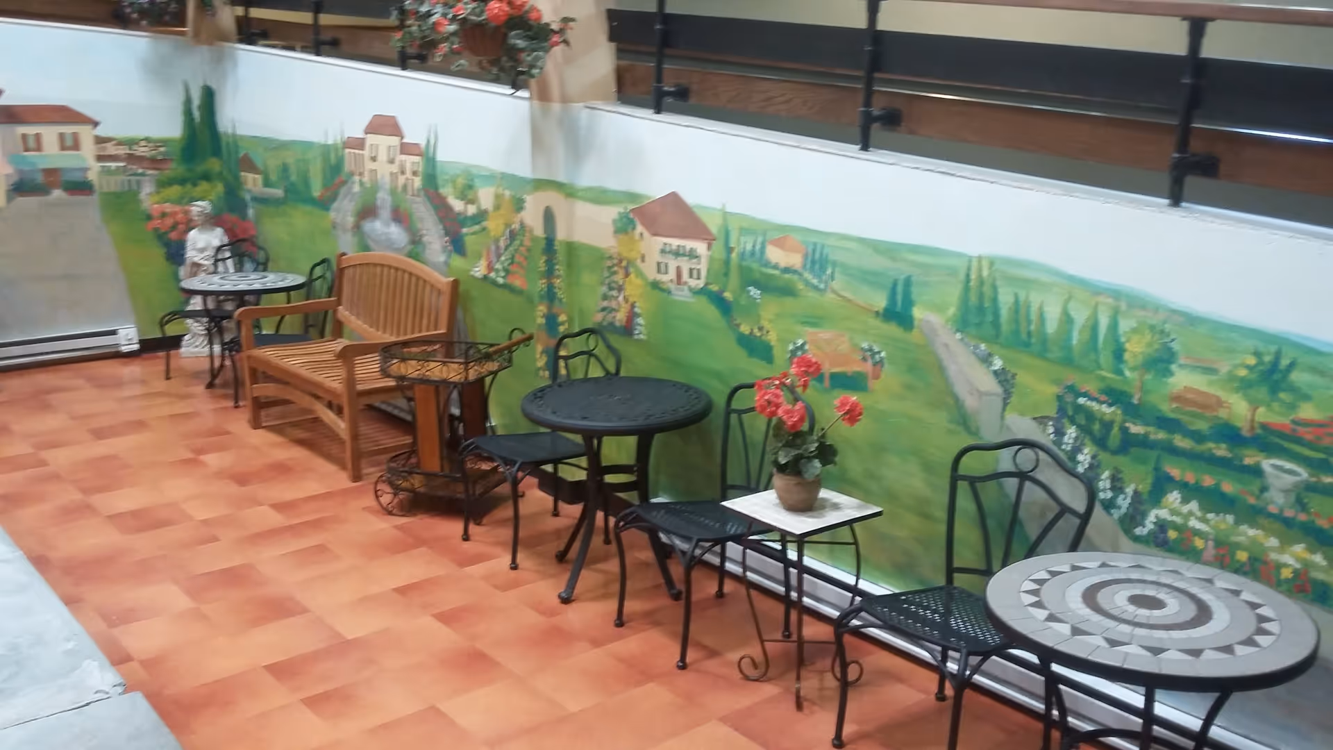 Indoor seating area with several small round tables and black metal chairs, a wooden bench, and a decorative statue. The wall behind features a colorful mural depicting a garden scene with houses, trees, flowers, and pathways.