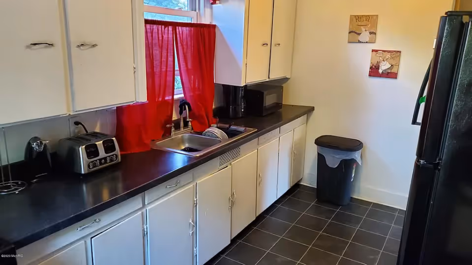A kitchen with white cabinets and black countertops. There is a stainless steel sink under a window with red curtains. On the counter, there is a toaster, a coffee maker, and a microwave. A black trash can is placed against the wall, and two decorative wall hangings are visible. The floor is covered with dark tiles.