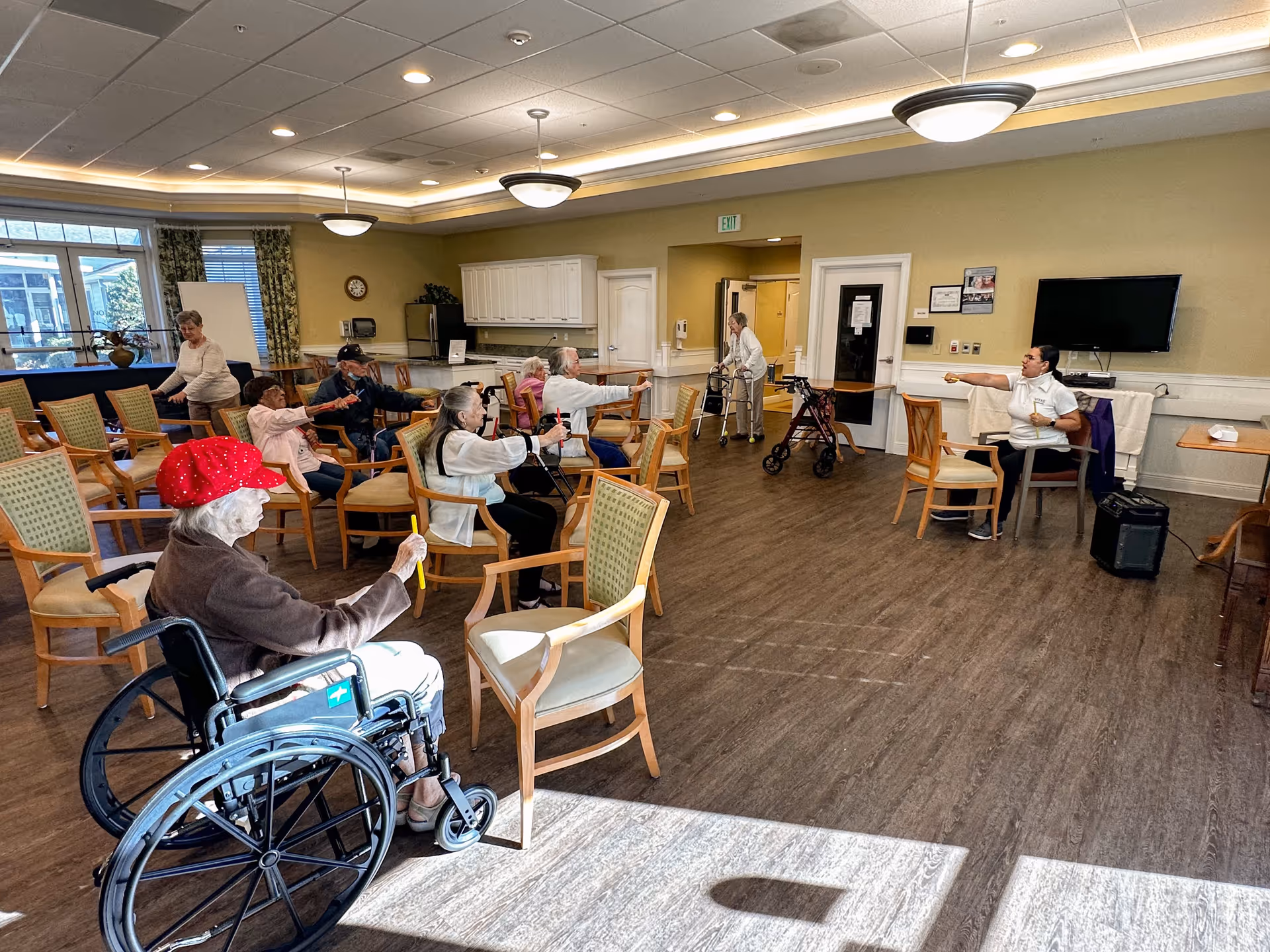 Residents in a senior living community sitting in chairs and a wheelchair doing a seated exercise class in a bright multi-purpose common room.