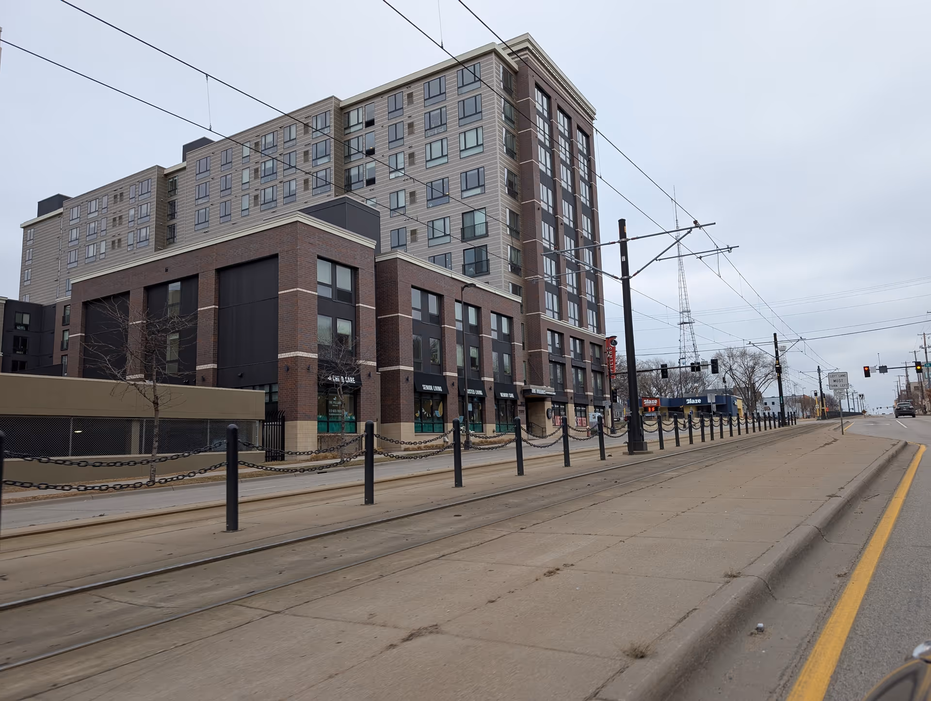 Multi-story brick-and-beige residential building with street-level shops beside tram tracks on an overcast day.