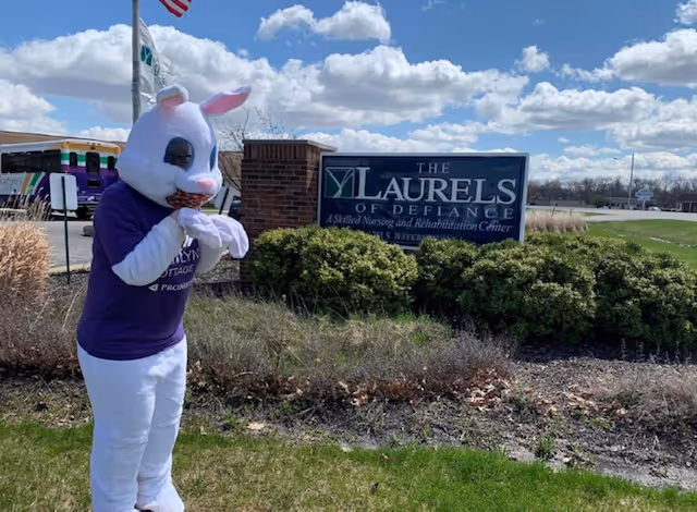 Person dressed in a white bunny costume with a purple shirt standing outside near a sign for The Laurels of Defiance, a skilled nursing and rehabilitation center. The scene is outdoors with grass, bushes, and a partly cloudy sky.