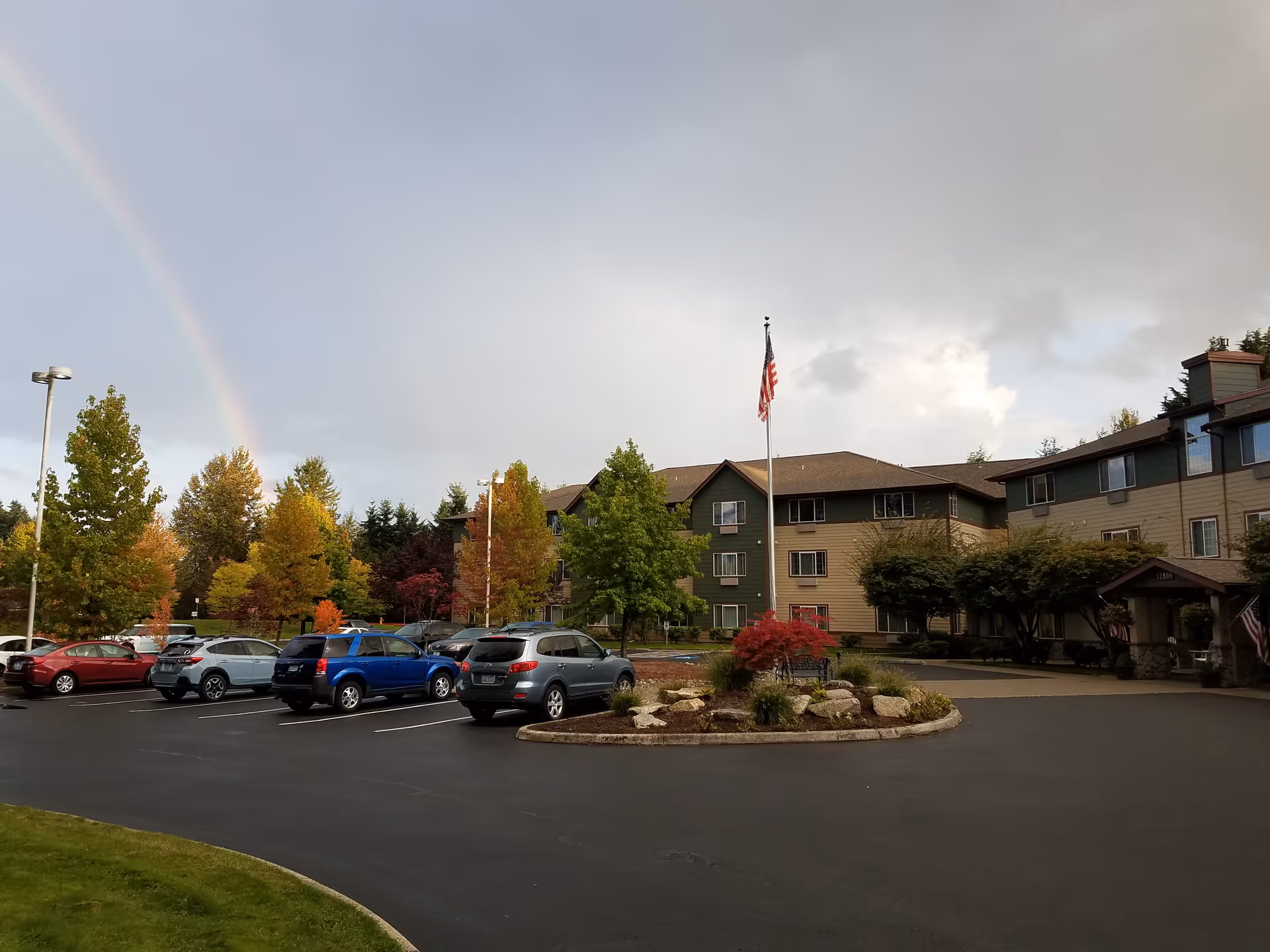 Exterior view of a senior living facility building with a parking lot in front. Several cars are parked, and there is a landscaped roundabout with rocks and plants in the center. An American flag is flying on a flagpole near the building entrance. Trees with autumn foliage surround the area, and a rainbow is visible in the sky.