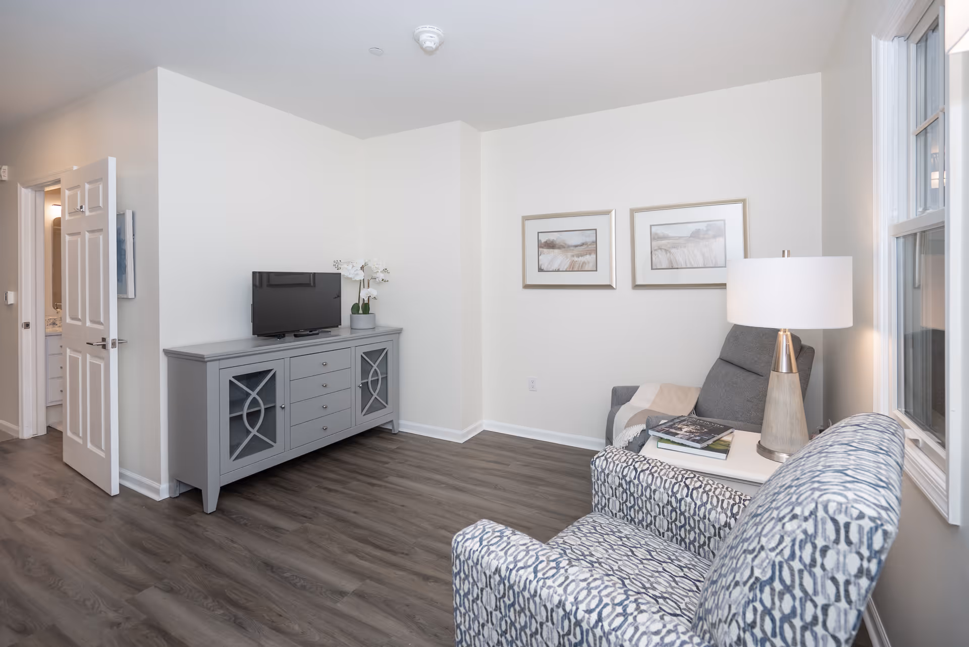 A cozy living room area in a senior living facility featuring a patterned armchair, a gray armchair with a throw blanket, a side table with a lamp and books, a gray TV console with a flat-screen TV and a white orchid plant, two framed landscape paintings on the wall, and a doorway leading to another room.