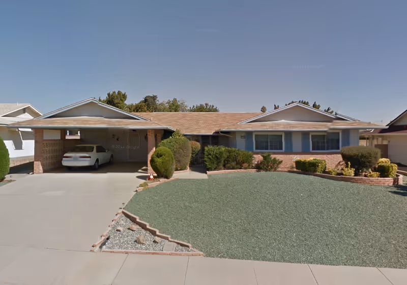 Single-story care home exterior with a carport, landscaped shrubs, and a wide driveway.