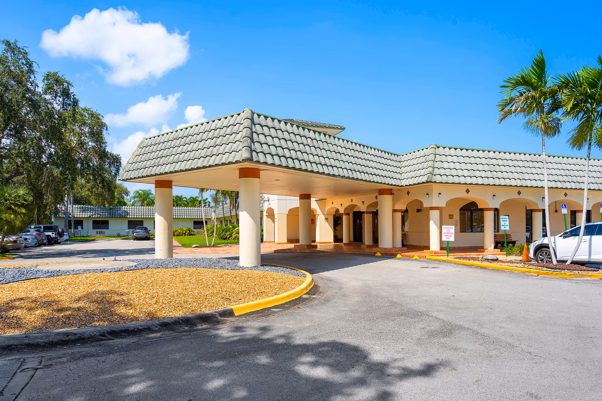 Exterior view of Shoreside Health and Rehabilitation Center showing the entrance with a covered drop-off area supported by columns, a tiled roof, palm trees, parked cars, and a clear blue sky.