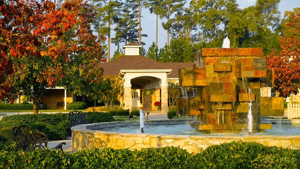 Outdoor scene at Atria Copeland featuring a large stone fountain with water flowing from multiple tiers, surrounded by green bushes and trees with autumn-colored leaves. In the background, there is a building with a covered entrance and a brown roof, set against a backdrop of tall pine trees.
