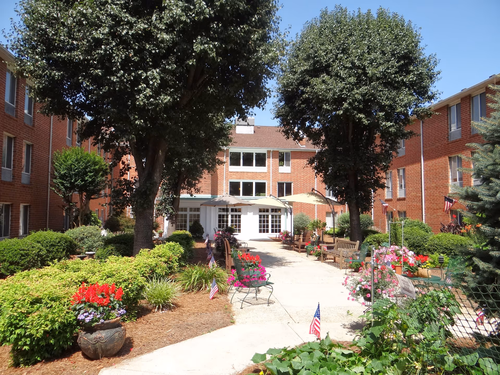 Outdoor courtyard area of Kingston Residence of Hickory with a paved walkway lined with benches, potted flowers, small American flags, and trees. The courtyard is surrounded by a red brick building under a clear blue sky.