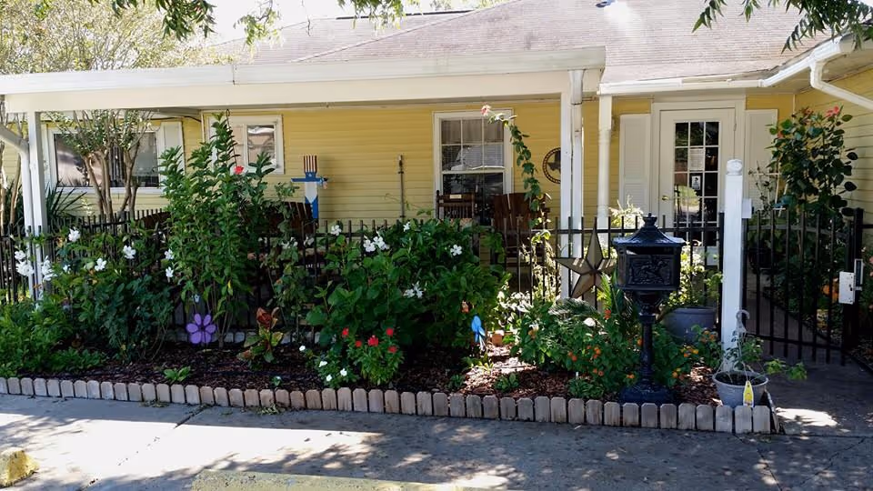 Front porch area of a yellow building with white trim, featuring a garden bed filled with green plants and flowers bordered by small bricks. There is a black mailbox on a post, a black metal fence, and a wooden star decoration. The porch has chairs and a door with glass panels.