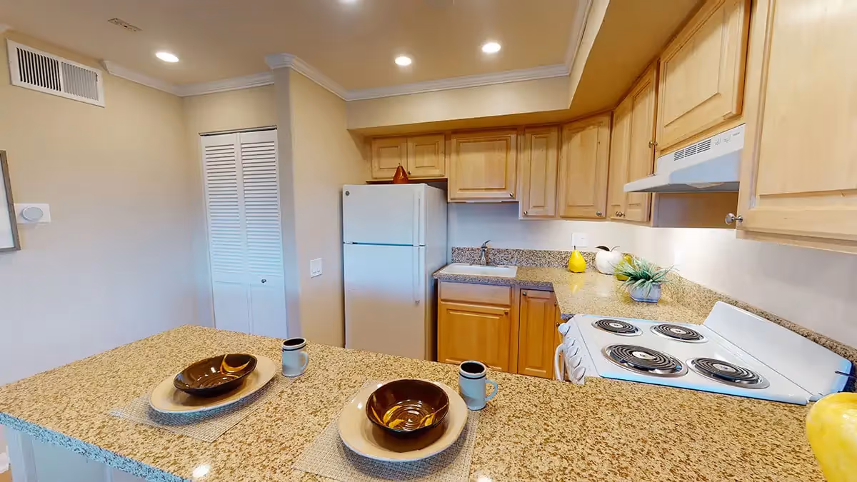 A kitchen area with light wood cabinets, a white refrigerator, a white electric stove with four burners, and a granite countertop with two place settings including plates, bowls, and mugs. There is a small sink under the cabinets and decorative items on the counter.