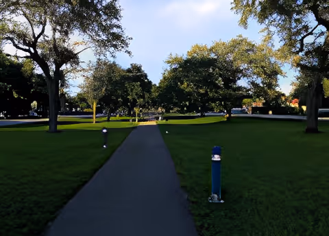 Paved walkway through a grassy, tree-lined lawn with small pathway lights under a partly cloudy sky.
