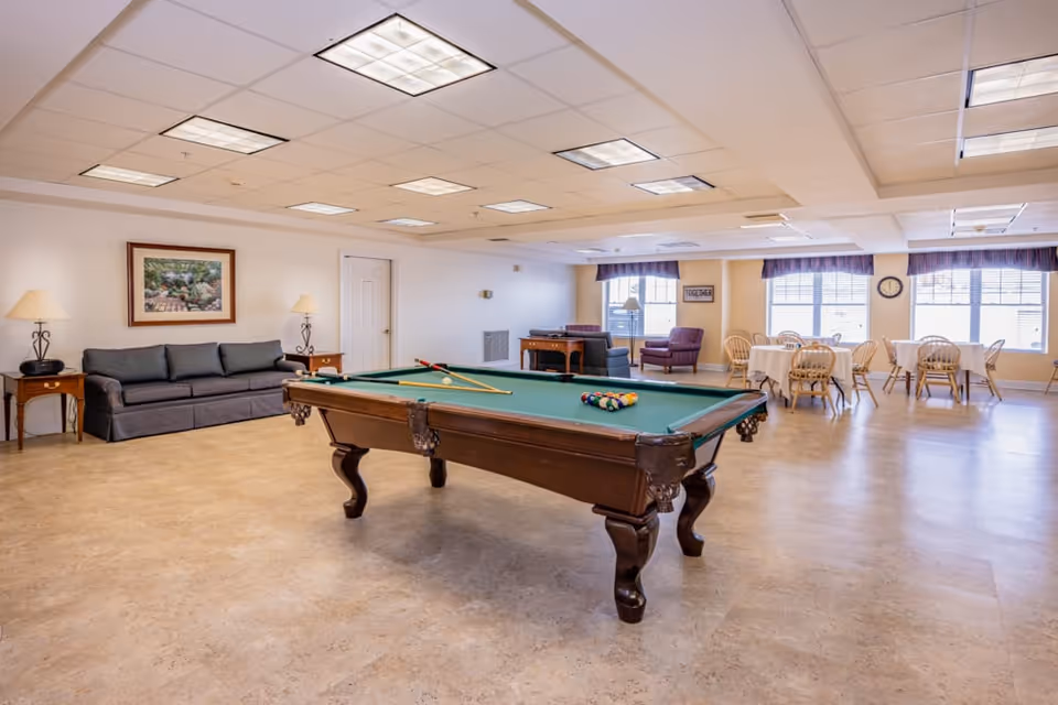 A spacious recreational room with a pool table in the center, a gray couch with side tables and lamps against the wall, and several round tables with chairs near large windows letting in natural light. The room has a light-colored floor and ceiling with recessed lighting.