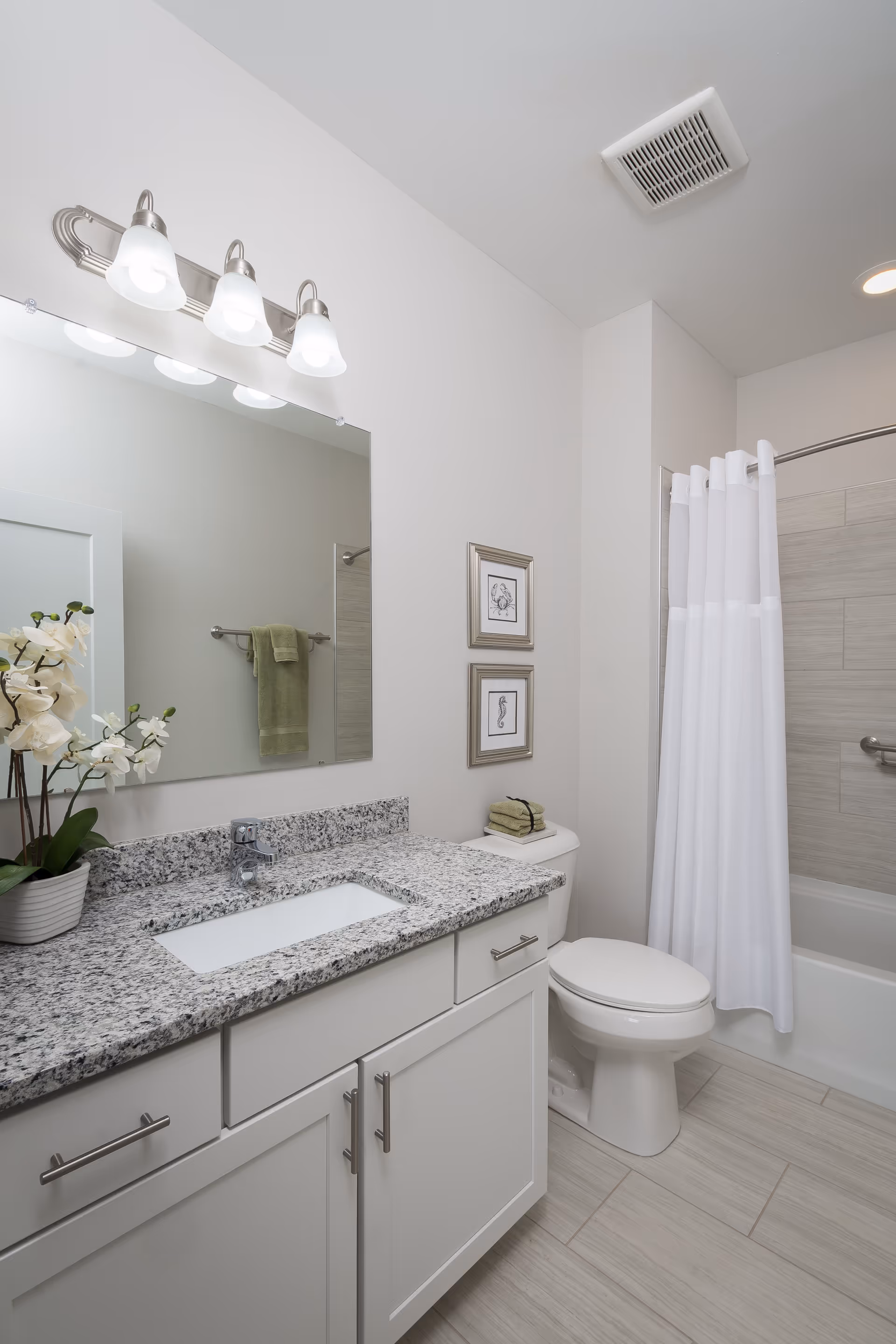 A clean and modern bathroom featuring a granite countertop with an under-mount sink, a large mirror with three light fixtures above it, a white toilet with neatly folded green towels on top, a white bathtub with a white shower curtain, light-colored tiled floor and walls, and two framed pictures on the wall.