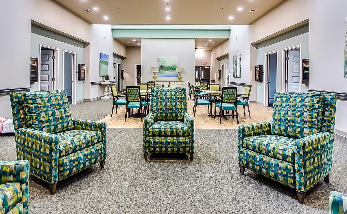 A spacious senior living common area with three patterned armchairs in the foreground and several dining tables with chairs in the background. The room has light-colored walls, artwork on the walls, and multiple doorways leading to other rooms.
