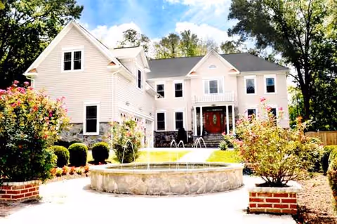 Two-story beige assisted living building with circular stone fountain and trimmed landscaping in front.