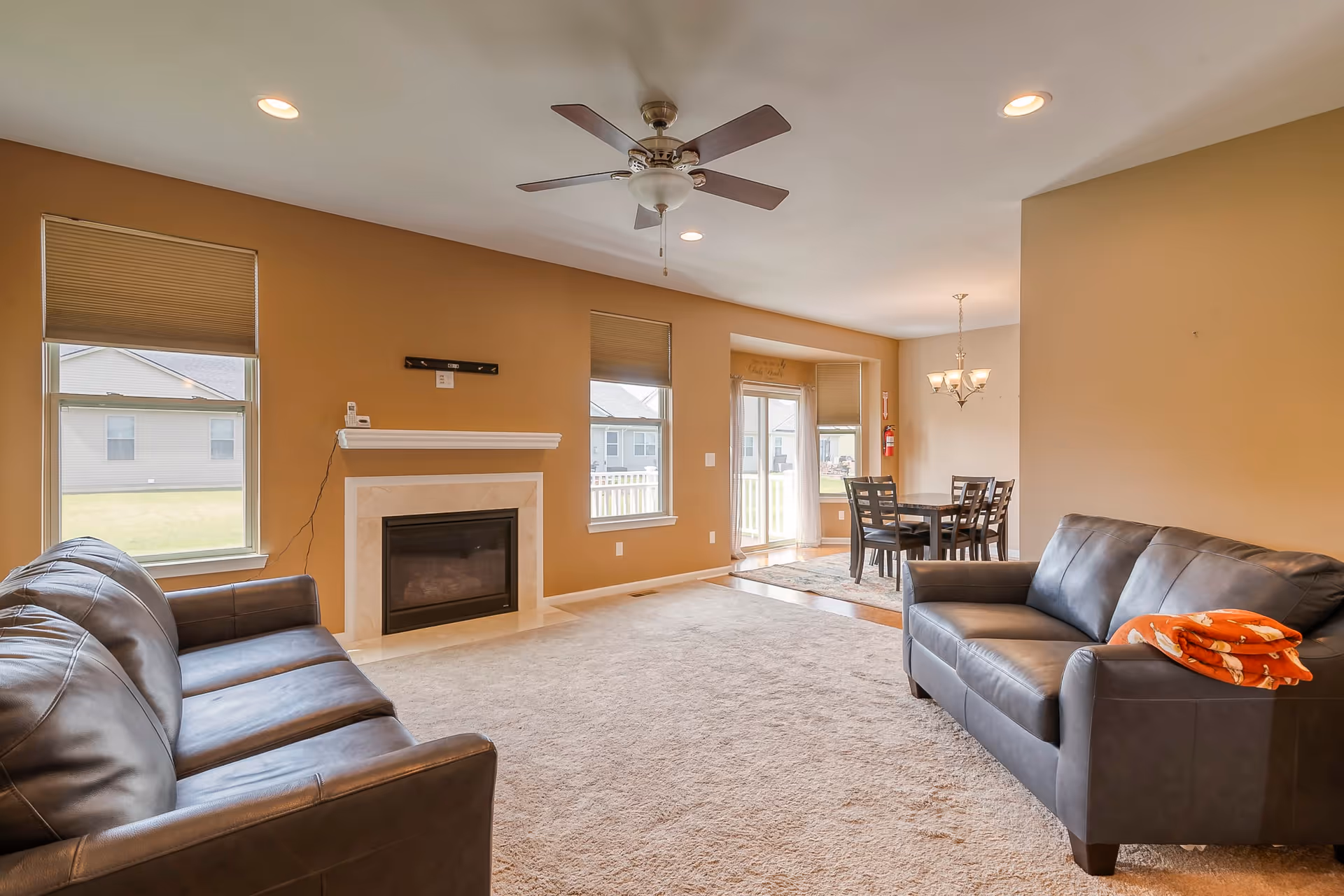 A spacious living room with two dark leather sofas facing each other, a beige carpet, a ceiling fan with lights, and a fireplace with a white mantel. The room has tan walls and two windows with blinds. In the background, there is a dining area with a wooden table and six chairs under a chandelier, next to glass doors leading outside.