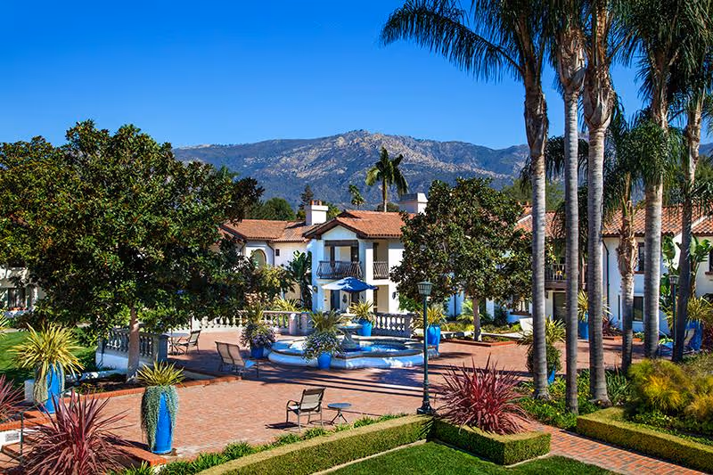 Outdoor courtyard area at Smith Health Care Center at Covenant Living at the Samarkand featuring a central fountain surrounded by trees, potted plants, chairs, and tables with a backdrop of mountains under a clear blue sky.