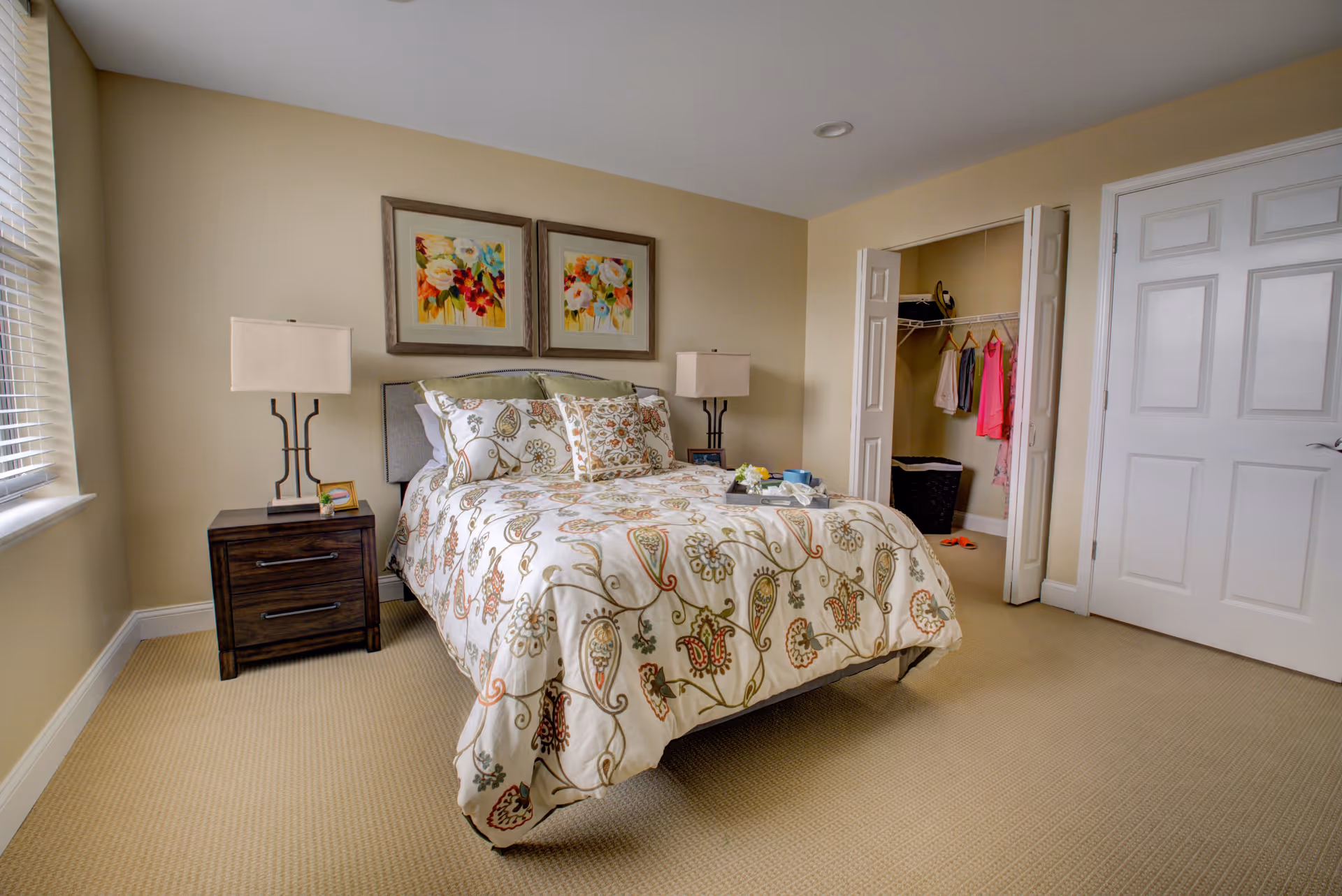 A cozy bedroom with a bed featuring a floral patterned comforter and multiple pillows. There are two framed floral paintings above the bed. On either side of the bed are dark wooden nightstands with matching lamps. To the right, there is an open closet with clothes hanging and a laundry basket inside. The room has beige walls and carpeted flooring, with a window on the left letting in natural light.