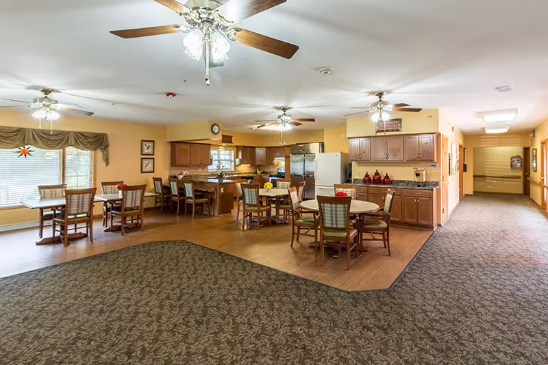 A spacious senior living facility common area featuring a kitchen with wooden cabinets, a refrigerator, and countertops. Several round and rectangular tables with chairs are arranged for dining or socializing. The room has ceiling fans with lights, large windows with valances, and a carpeted floor transitioning to wood near the kitchen area. A hallway extends from the right side of the room.